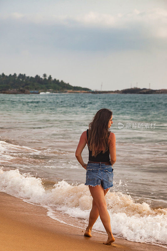 后视图年轻女子在牛仔短裤走在沙滩上的海浪背景，暑假。后视图女士走在热带海岸，生活方式。旅游度假概念。复制文本空格