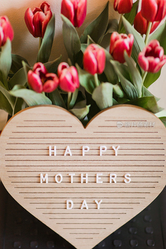 一个心形的字母板，上面写着“母亲节快乐”，明亮的粉红色郁金香盛开，郁郁葱葱的绿色茎和叶子在它后面，在明亮的自然日光下拍摄