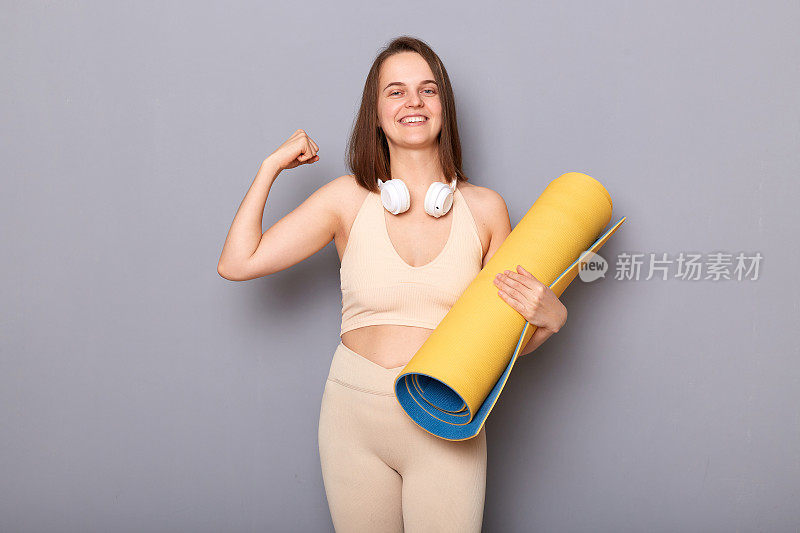 满意的高兴的女人的肖像棕色头发站在瑜伽垫和耳机在脖子上，显示她的肱二头肌，微笑，享受锻炼和减肥的结果。
