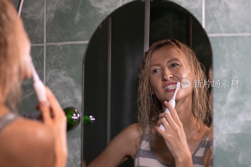 年轻的金发女郎正在浴室里用一把白色电动牙刷刷牙，看着镜子。卫生，身体护理的理念。