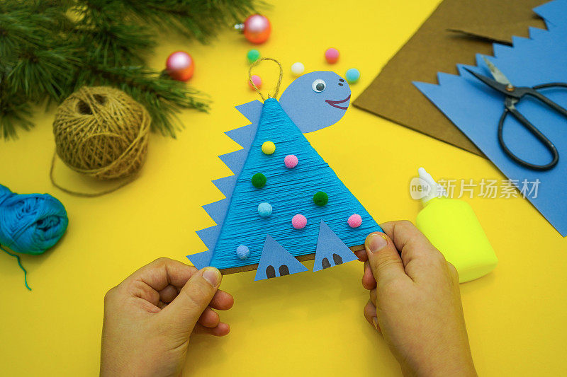 手和圣诞树的特写:一个孩子用硬纸板和红线做的恐龙玩具躺在黄色的背景上。2024年的象征。教育和手工艺活动。