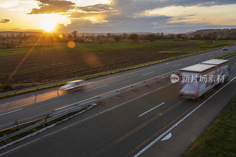 一辆载着两辆拖车和两辆汽车的大卡车在公路上高速行驶，穿过乡村风景。高速公路高速行驶。高速公路上超速行驶的景象。背景是美丽的日落。