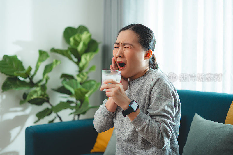 亚洲女性坐在客厅的沙发上，喝了凉水，摸了摸脸颊，就会感到牙齿敏感。