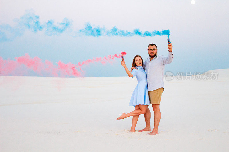 年轻的情侣站在沙滩上，手里拿着彩色烟雾弹，浪漫的情侣带着蓝色和红色的烟雾弹在沙滩上