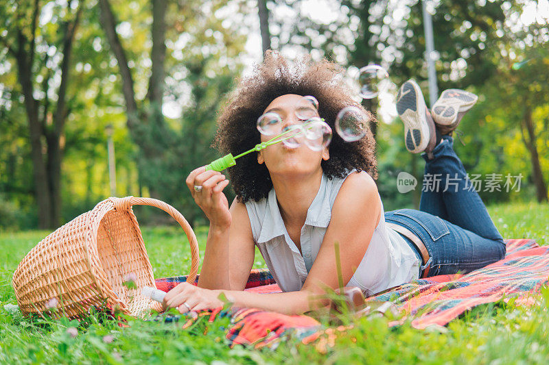 一个年轻的女人躺在户外的毯子上制造肥皂泡