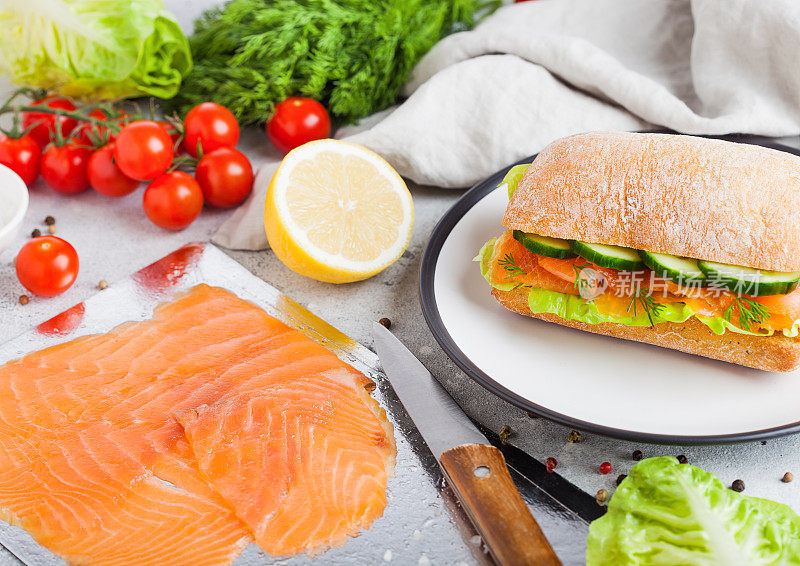 新鲜健康的三文鱼三明治与生菜和黄瓜的盘子在白色的石头背景。早餐吃零食。新鲜番茄，莳萝和柠檬。用刀和盘子盛鲑鱼