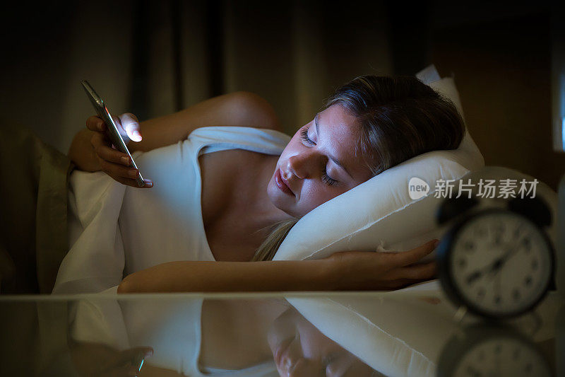 年轻漂亮的女人晚上在卧室里用智能手机醒来。滥用手机的概念。