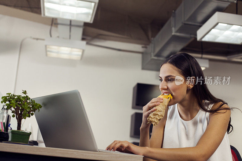 一位拉丁妇女，在电脑前专心工作，一边快速吃着三明治