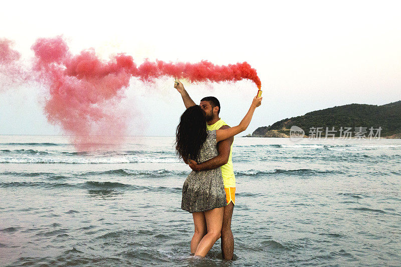 年轻的情侣站在沙滩上，手拿烟雾弹，看着对方，浪漫的情侣带着黄色和红色的烟雾弹在沙滩上