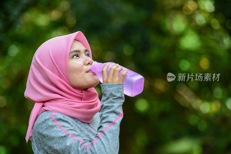 年轻的亚洲穆斯林女运动员戴着头巾在慢跑的跑道上喝着水瓶里的水