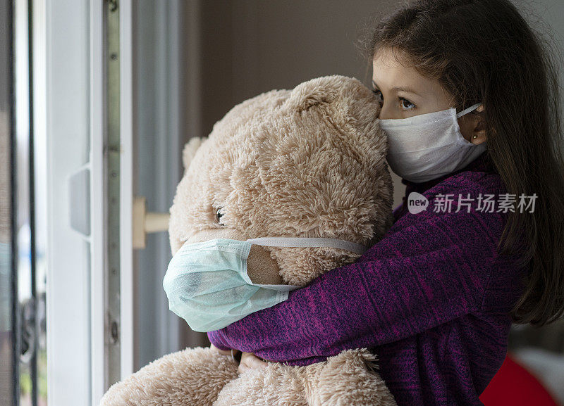 带着防护面具的可爱小女孩和她的泰迪熊站在窗边