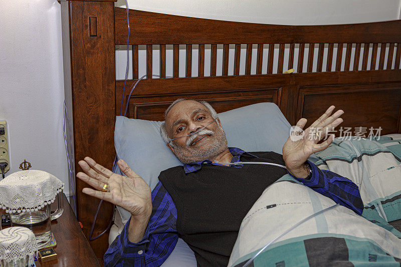 一名72岁的印度亚裔男子在接受腹腔镜胆囊手术后正在家中的床上康复。他在镜头前做了一个手势，表示他克服了困难。