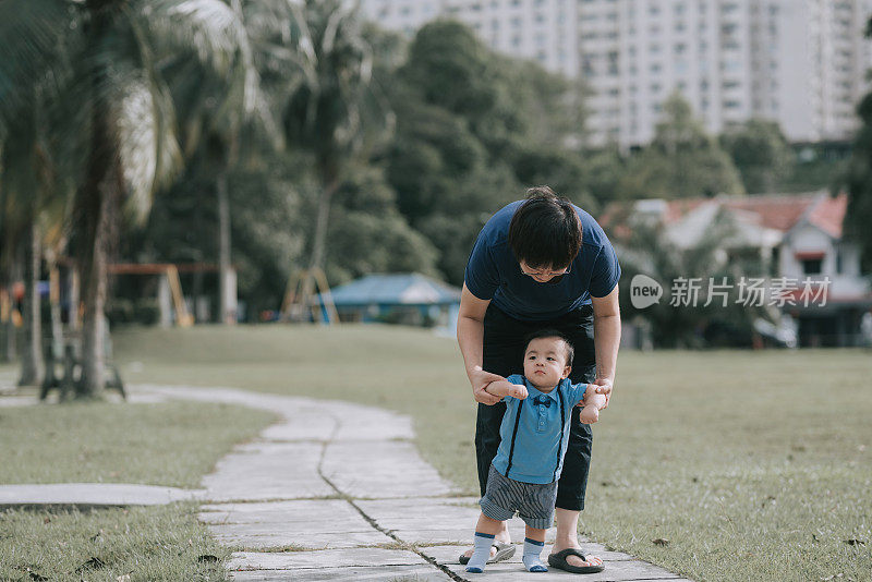 亚洲华人母亲牵着他的儿子的手，教他，并鼓励他早上在公园学习的第一步