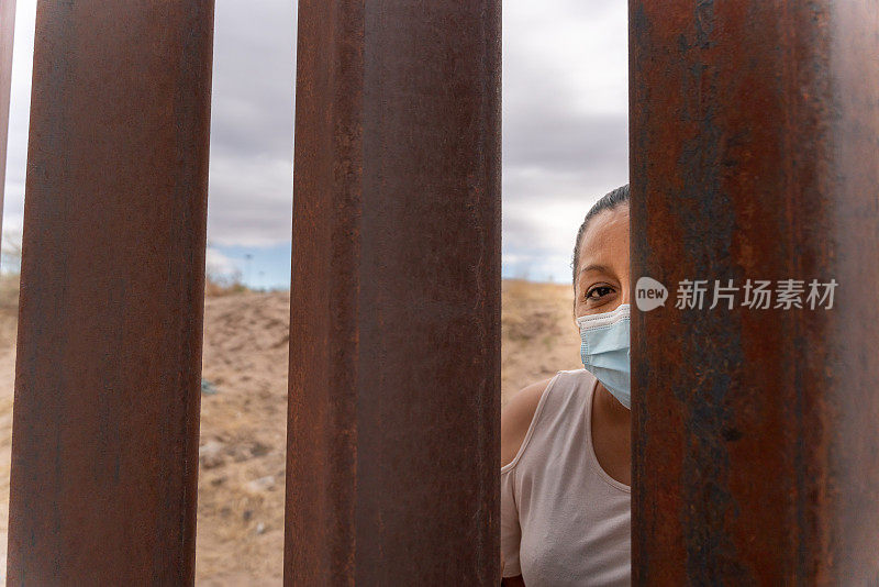 中年成年墨西哥妇女戴着防护面具，在美国和墨西哥之间的国际边境隔离墙后摆姿势
