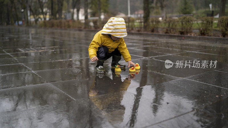 可爱调皮的小男孩，穿着亮黄色雨衣和胶靴，在春雨天潮湿的街道上和小水坑里的橡皮鸭玩耍。季节性天气步行概念