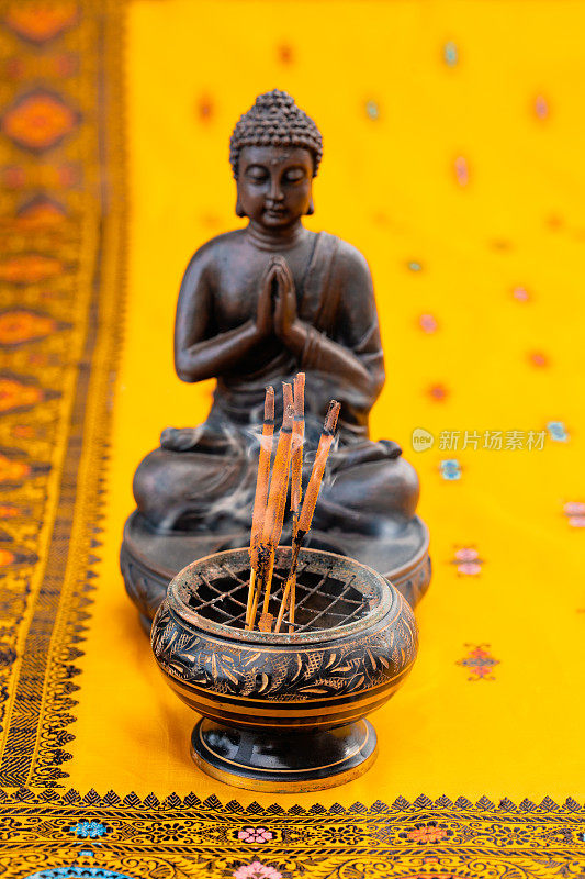 在一个没有聚焦的棕色佛像前的香碗里熏香，还有带有刺绣的黄橙色印度教纱丽绳