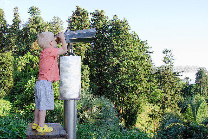 金发小男孩在看一副大望远镜。背景中的公园