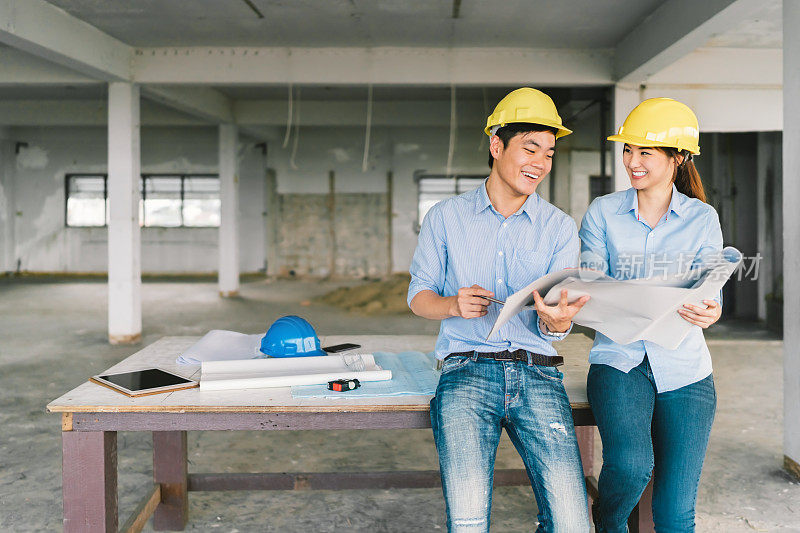 年轻的亚洲工程师夫妇一起工作在建筑工地或工厂的建筑蓝图。土木工程，工业商业伙伴，或家居装修服务理念。与复制空间