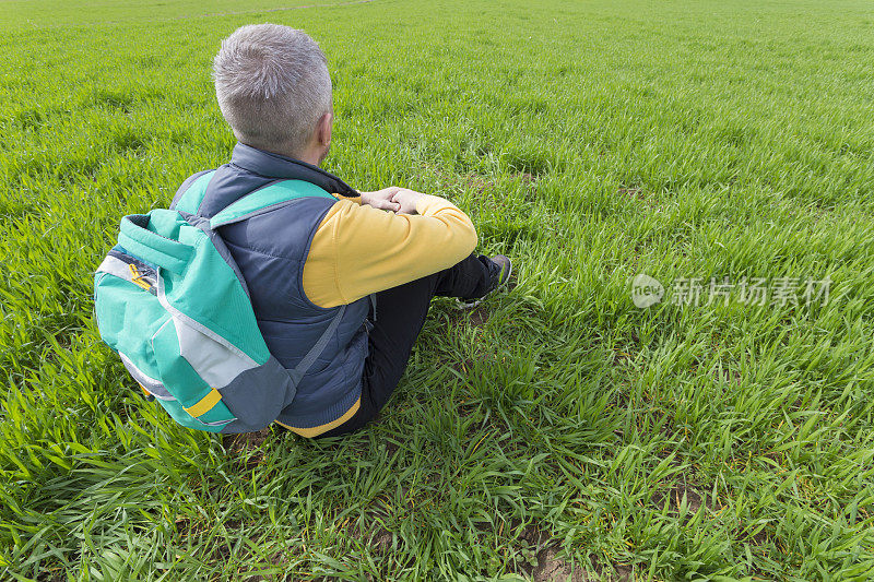 一名成年男子背着双肩包徒步旅行，坐在草地上休息，这是初春阳光明媚的一天。后面的观点。