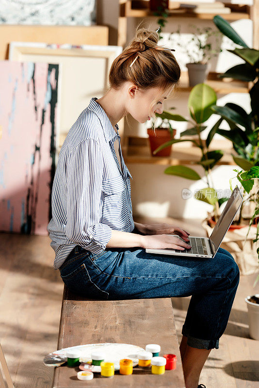 侧面的年轻女性艺术家坐在长凳上与颜料和使用笔记本电脑