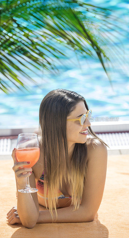 美丽的年轻女子日光浴在比基尼附近的游泳池喝鸡尾酒