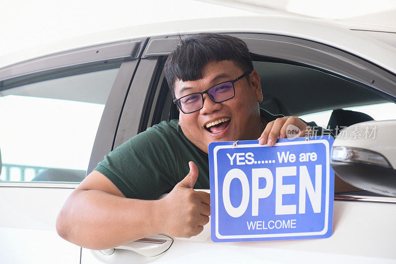 一名亚裔男子坐在车里，举着“开放标志”的海报，竖起大拇指。