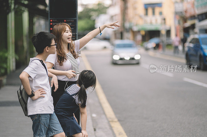 一位亚洲华人妈妈带着她的儿子和女儿旅行，在路边用手机应用拦了一辆出租车，并向司机挥手