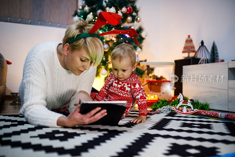 冬日里，在温馨的客厅里，年轻的母亲和她快乐的小男孩在圣诞树旁的圣诞背景上放着数码平板电脑