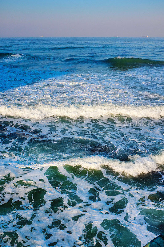 海浪从海洋中滚滚而来，形成泡沫状的浪花，远处的船只出现在地平线上