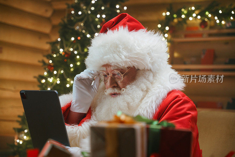 圣诞老人坐在挂着圣诞灯的小木屋里，用礼物装饰圣诞树，全神贯注地看着数码平板电脑