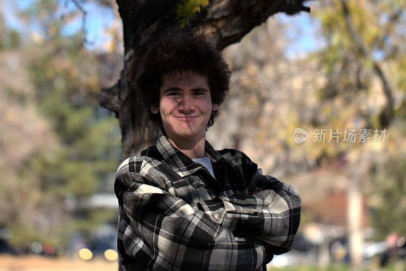 年轻，微笑，卷发的白人男孩站在一棵树前，双臂交叉
