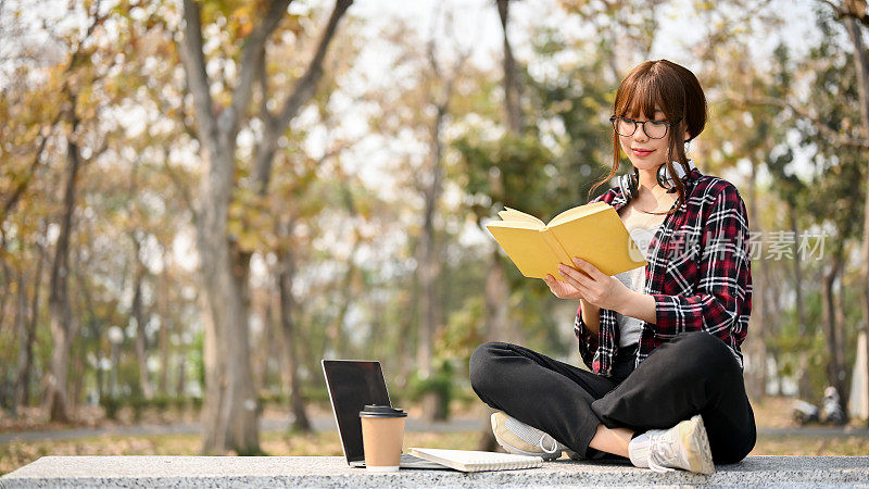 漂亮的亚洲女学生专注于在校园公园的长椅上看书。