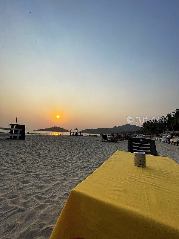 海滩餐厅设置的特写图片，桌子和椅子准备在日落时露天用餐，黄色桌布覆盖的桌子，印度果阿的Palolem海滩
