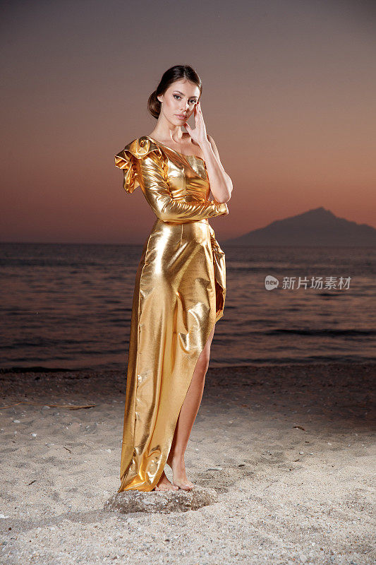 美丽的年轻女子在沙滩上的金色长裙黄昏