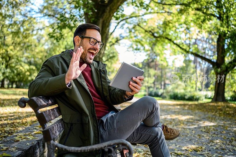 一个面带微笑的男人坐在公园的长椅上，用着一台数码平板电脑