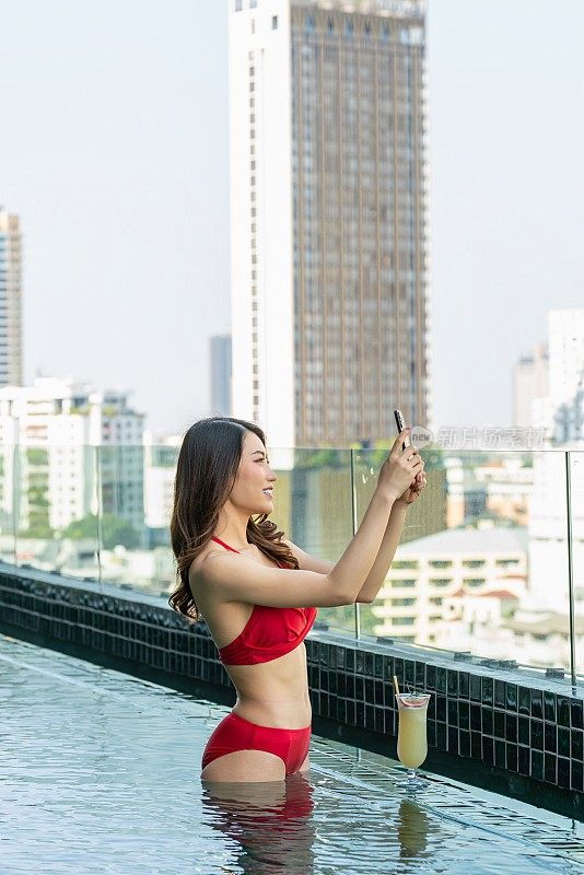 城市风光自拍:女子开心地笑着，在屋顶泳池边用手机拍下这一刻，旁边有饮料，背景是城市风光。