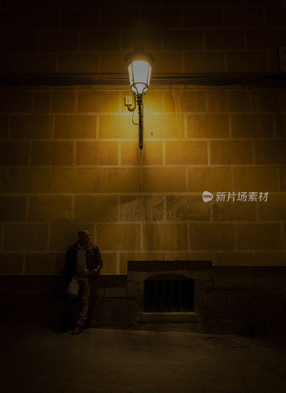 成年男子在街上靠砖墙，路灯在夜间。
