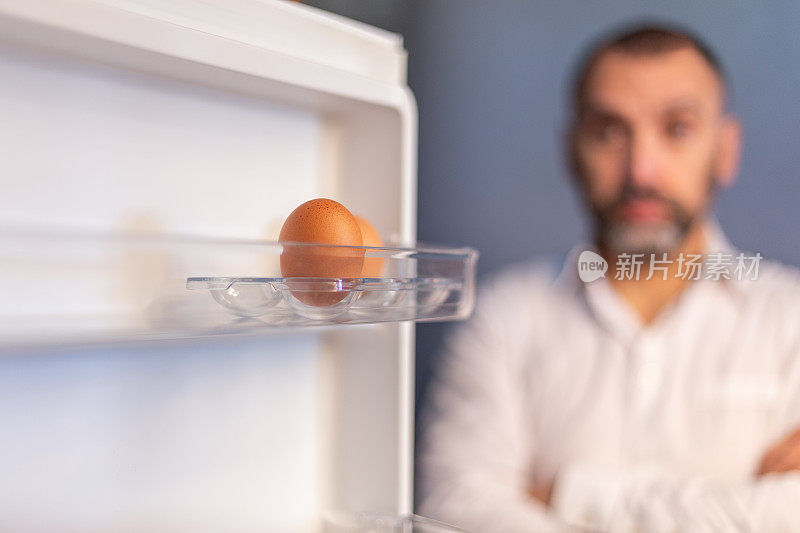 冰箱门上有两个鸡蛋。背景中，一名心不在焉的男子盯着空无一物的冰箱。概念:空冰箱。没有吃的吗?