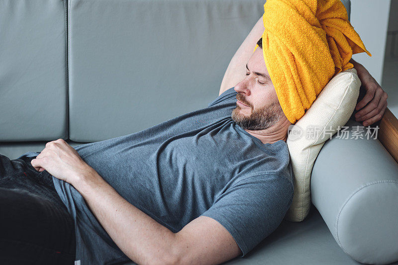 帅哥躺在沙发上，眼睛上放着黄瓜片，头上放着黄色的毛巾。男性自我护理理念，护肤，在家放松。
