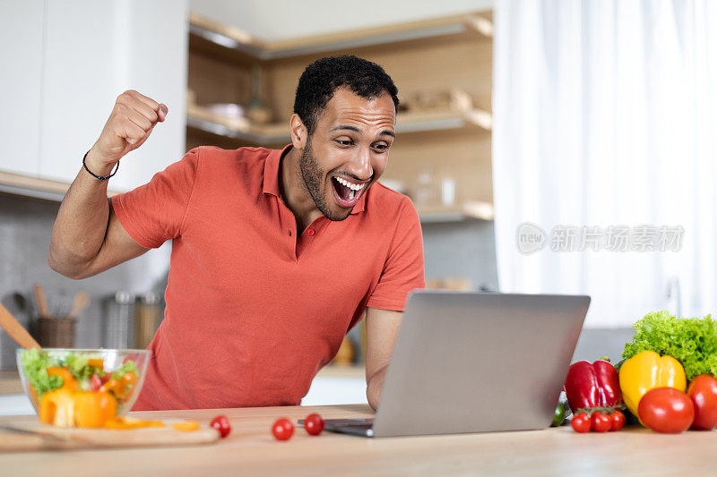 笑兴奋的千禧一代非洲裔美国人在桌子上有机蔬菜使用笔记本电脑，使成功的姿态