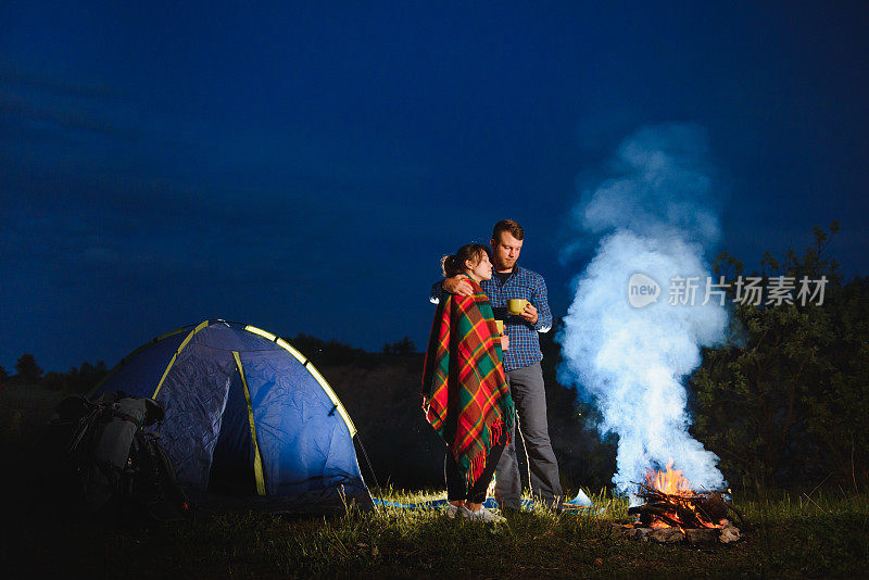 爱的夫妇徒步旅行彼此享受，站在篝火篝火在傍晚的天空下，靠近树木和帐篷。在山上的森林附近浪漫露营