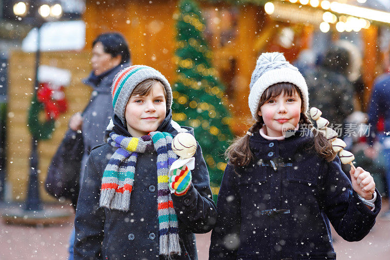 可爱的小女孩和男孩在传统的德国圣诞市场上吃着白巧克力包裹的草莓和苹果串。快乐的孩子，最好的朋友，双胞胎和兄弟姐妹在雪天