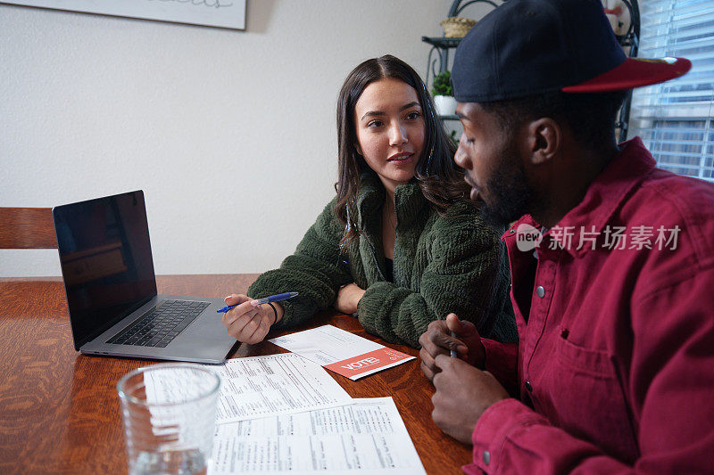 年轻的混血夫妇西班牙裔和非裔美国人坐在厨房的桌子上研究问题和候选人，填写邮寄投票