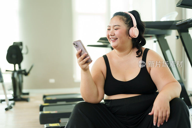 大码的亚洲女性在健身房锻炼，穿着运动服的女性在休息时愉快地使用智能手机，体现了休闲和健身的平衡。舒舒服服地坐在跑步机上，快乐就会到来