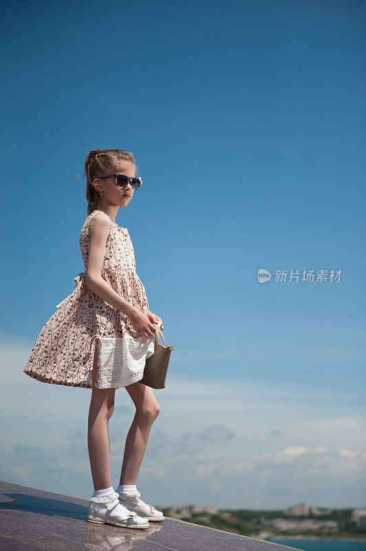 时尚的小女孩穿着时髦的衣服，戴着太阳镜，拿着小袋子站在蓝天的背景上
