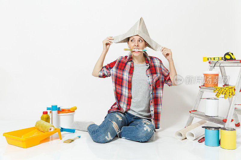 戴着报纸帽子的女人坐在地板上，嘴里叼着刷子，仪器为装修公寓孤立在白色的背景上。墙纸，胶水配件，油漆工具。修理家里的概念。