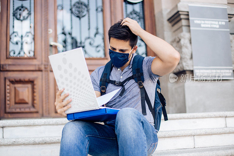 一个戴着口罩的学生坐在他的大学前面读书的肖像