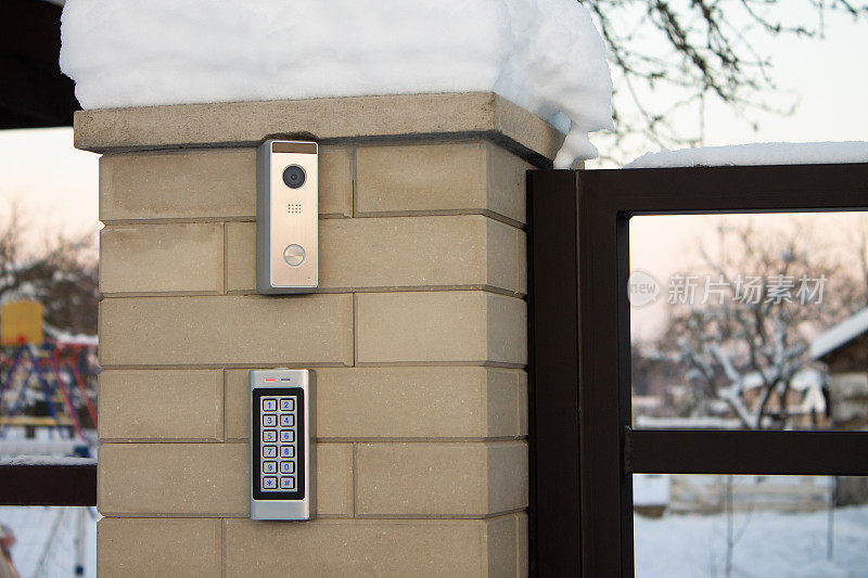 银色对讲机呼叫面板，蓝色数字按钮和一个摄像机在一个私人住宅的砖米色围栏柱子上