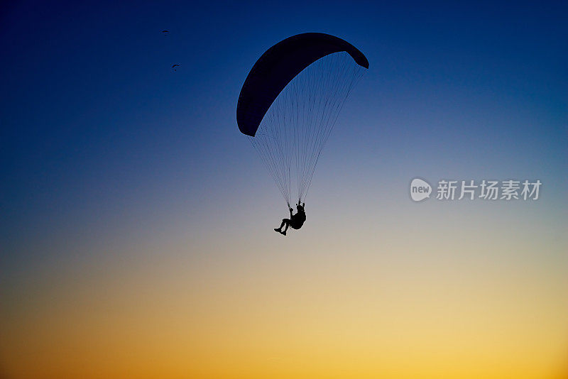 黑色剪影的滑翔伞飞行在晴朗的傍晚蓝黄色的天空背景