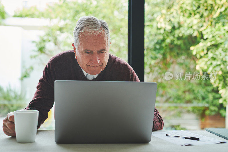 照片中，一位老人独自坐在家里，一边用着笔记本电脑，一边喝着咖啡